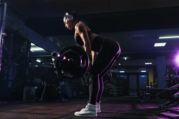 Attractive fit sexy woman in wireless headphones listen music and doing crouches with a barbell in the gym. woman training back