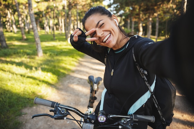 Attraente sportiva in forma in sella a una bicicletta al parco, ascoltando musica con auricolari wireless, prendendo un selfie