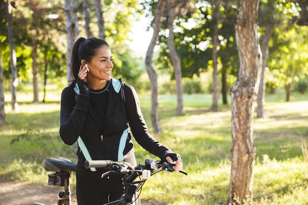 Привлекательная спортивная женщина с велосипедом в парке, слушает музыку в беспроводных наушниках