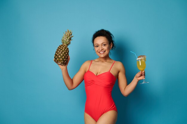 Attractive fit woman in red swimsuit posing  with a glass of cocktail and a pineapple in her hands