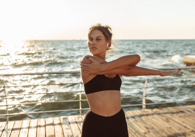 Привлекательная женщина в спортивной одежде делает растяжку рук перед тренировкой на пляже на рассвете