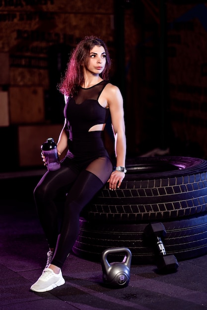 Attractive fit woman with shaker posing on big tire in the gym.