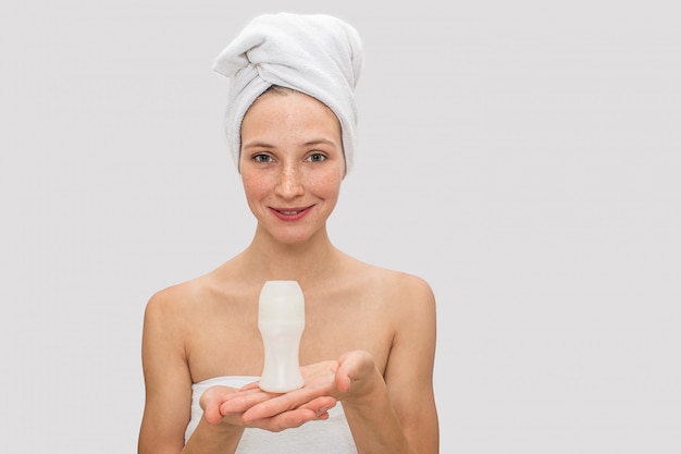 Attractive and freckled woman poses. She holds deodorant with both hands. She smiles. There are white towel around her hair.