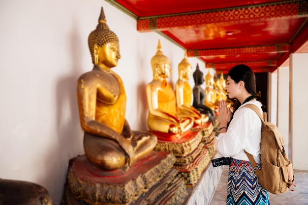 仏の前に立って、タイ旅行のバンコクのワットポー寺院で健康的な幸福を願うジェスチャーを祈る魅力的なフレンドリーな女子学生。