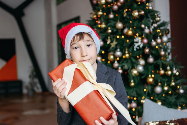 Attractive funny tween boy with dark hair in santa hat with gift box on christmas tree 