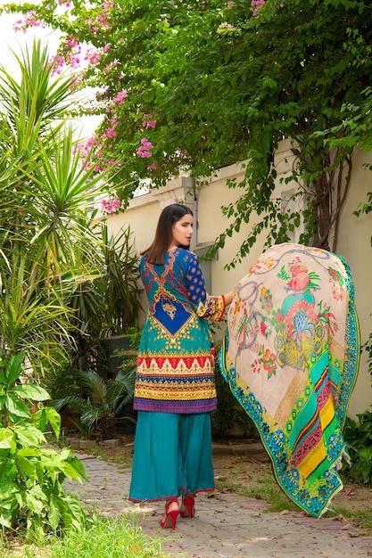 Attractive Girl Back Pose Waving Dupatta in House Garden with Plants Wearing Trendy Desi Dress