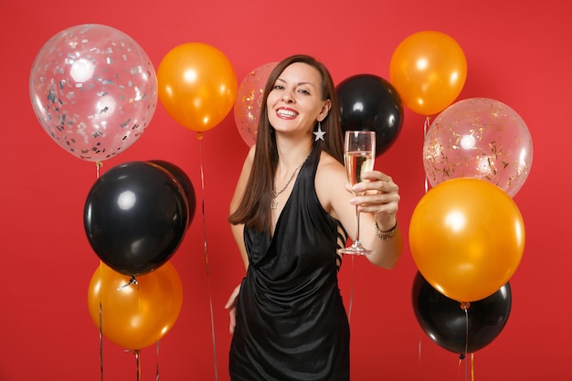 Attractive girl in black dress celebrate, pointing hand with glass of champagne on camera on bright red background air balloons. Valentine's Day, Happy New Year, birthday mockup holiday party concept.