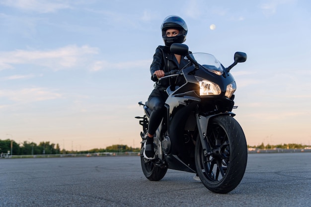 Attractive girl in black leather jacket pants and helmet on outdoors parking rides on stylish