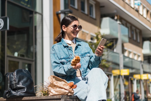 Attractive girl eating in the street in the city while using phone to rate the takeaway food company for delicious menu