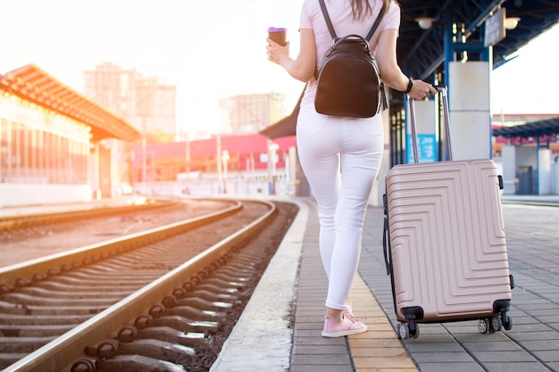 Attractive girl is standing with luggage at the station and waiting for the train, the student is going on a trip, she is walking along the platform with coffee, copy space