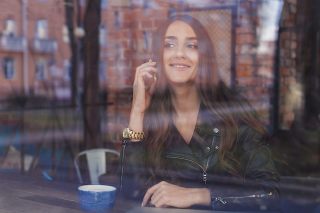 An attractive girl in a leather jacket talking on a mobile phone and smiling outside a cafe.