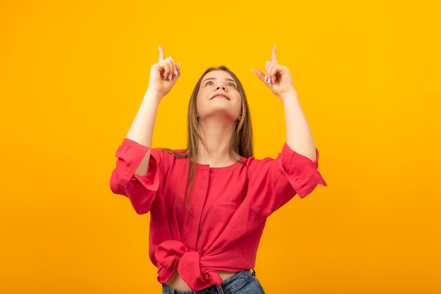 Attractive girl looks up and points her fingers at something Young smiling woman pointing up yellow background