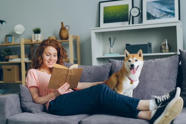 Attractive girl reading book and stroking shiba inu puppy on couch in flat