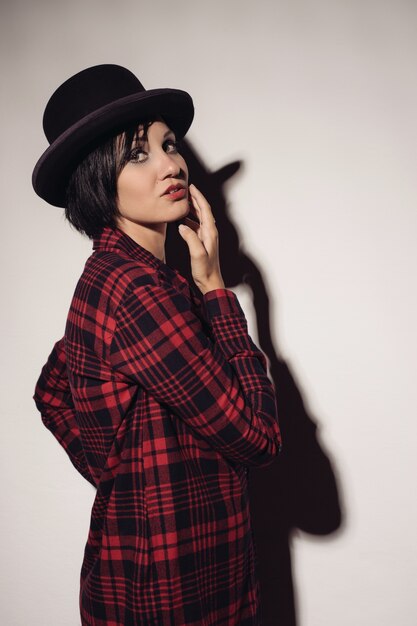 Attractive girl in red checkered shirt and hat bowler stands near wall raising her hand to face