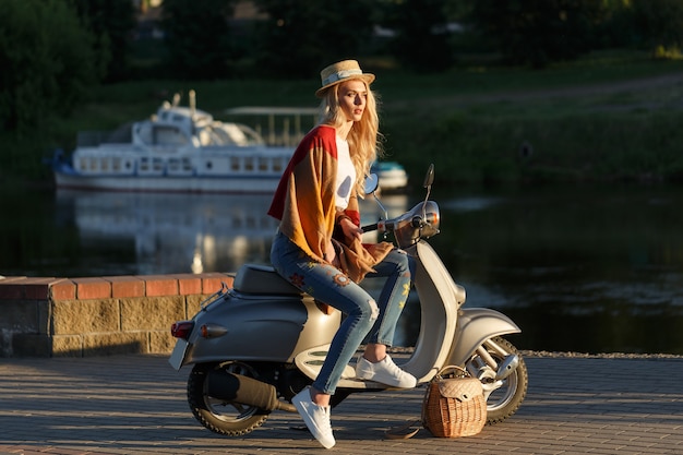 매력적인 여자의 초상화입니다. 해질녘 강 근처의 복고풍 오토바이에 앉아 청바지, 짧은 티셔츠, 밀짚모자를 입은 여성. 여행 개념입니다.