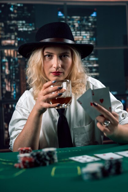 Foto attraente ragazza in camicia e cappello che beve whisky e guarda le carte da poker nel casinò una ragazza giocatrice fa una scommessa poker nel casinò