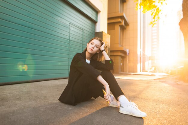 Attractive girl sitting on asphalt background of green wall and sunset