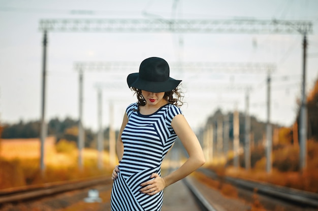 Photo attractive girl in a striped dress and hat is walking on the railroad