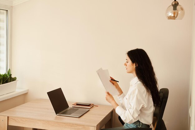 Attractive girl student studying online using laptop at home Online education and distance learning Girl holding papers in hands