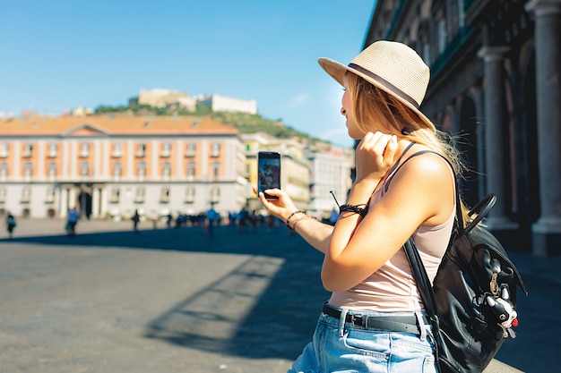 Attractive girl tourist in hat with backpack exploring new city in europe at summer and using her phone to take photo