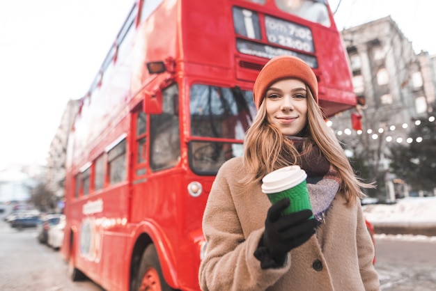 Attraente ragazza in caldi abiti primaverili, in piedi con un bicchiere di carta di caffè nelle mani di un autobus turistico rosso, guardando la telecamera e sorridente