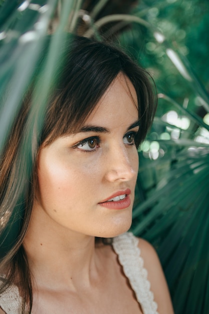 Attractive girl with brown long hair posing in front of green trees while touching the branches