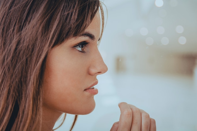 Attractive girl with brown long hair posing near the window with her hand on the glass