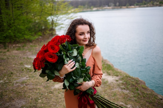 Attraente ragazza con i capelli ricci e un sorriso sul viso con un enorme mazzo di rose rosse su uno sfondo di lago blu. calda giornata estiva, giovane donna felice, emozioni di gioia