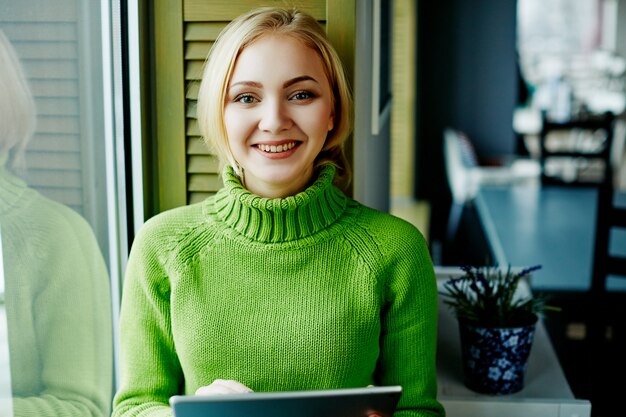 Привлекательная девушка с светлыми волосами в зеленом свитере, сидя в кафе с планшетом, внештатная концепция, интернет-магазины, портрет.