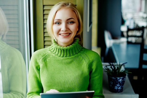 Attractive girl with light hair wearing green sweater sitting in cafe with tablet, freelance concept, online shopping, portrait.