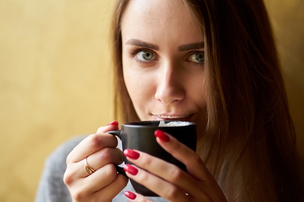 Привлекательная девушка с длинными волосами и губами в пене держит чашку капучино и пьет кофе