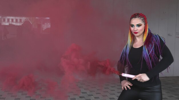 Привлекательная девушка с разноцветными косами и макияжем в черном платье. Позирует, скрываясь за красным искусственным дымом на фоне здания в весеннем городе.