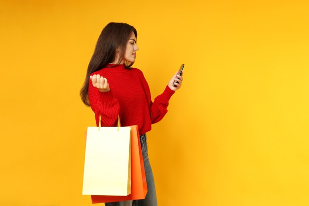 Attractive girl with paper bags and smartphone on yellow background