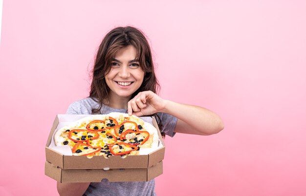 Attractive girl with pizza in a box for delivery on a pink background