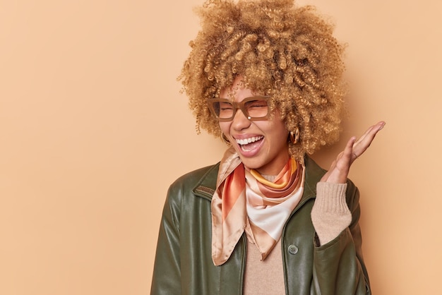 Attractive glad young woman exclaims happily keeps hand raised closes eyes from joy wears spectacles leather jacket and kerchief around neck reacts on awesome news poses against beige background