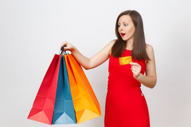 Attractive glamour fashionable young brown-hair woman in red dress holding credit card, multi colored packets with purchases after shopping isolated on white background. Copy space for advertisement.