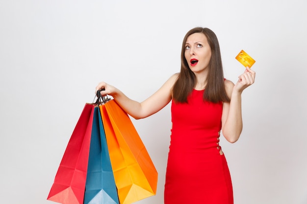 Attractive glamour fashionable young brown-hair woman in red dress holding credit card, multi colored packets with purchases after shopping isolated on white background. Copy space for advertisement.