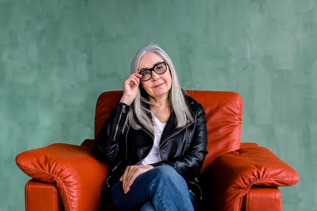 Attractive gray haired lady in black leather jacket and fashionable eyeglasses, sitting in red armchair on green background, looking at camera with smile