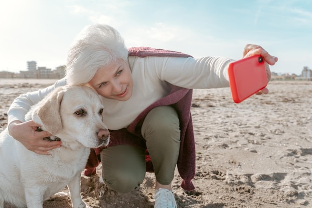 Attractive gray-haired lady sitting on the haunches and taking a selfie with her obedient pet