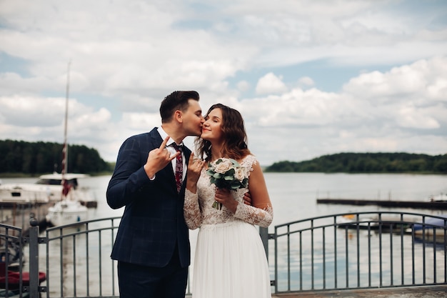 Sposo attraente in abito nero con la sua bella sposa in abito bianco lungo vicino al lago