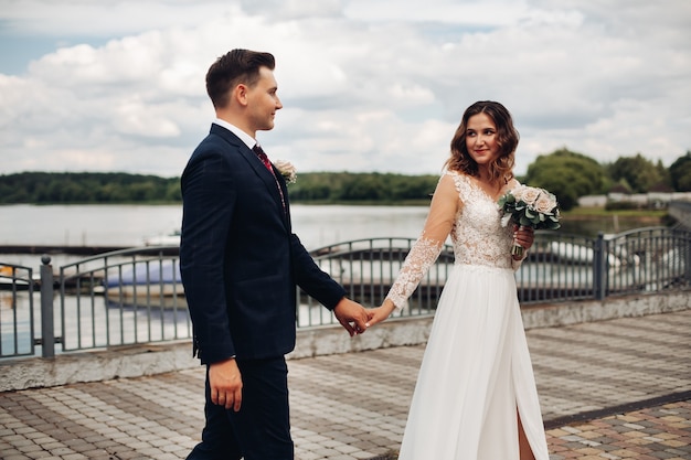 Attractive groom in black suit with his beautiful bride in long white dress near the lake