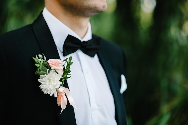 The attractive groom with bow tie in a suit with boutonniere or\
buttonhole on jacket is stands on the background greenery in the\
garden park nature cropped photo