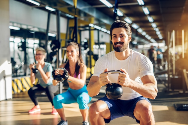 매력적인 잘 생긴 수염 난된 남자가 kettlebell을 들고 현대 체육관에서 두 여자와 피트니스 그룹에서 스쿼트를하고 있습니다.