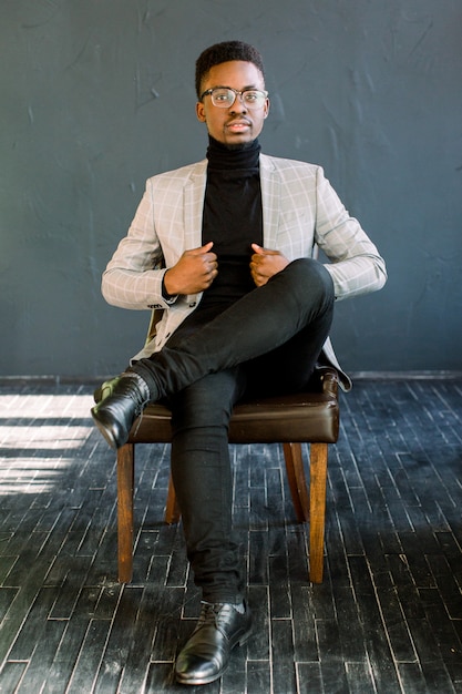 Attractive handsome happy smiling African American businessman wearing a light jacket and glasses sitting on a leather chair on a dark background