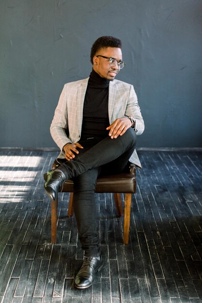 Attractive handsome happy smiling African American businessman wearing a light jacket and glasses sitting on a leather chair on a dark background