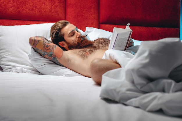 Attractive handsome nice man with a mustache and a beard lying on the bed and reading a book before dreams in a hotel room. Night time concept