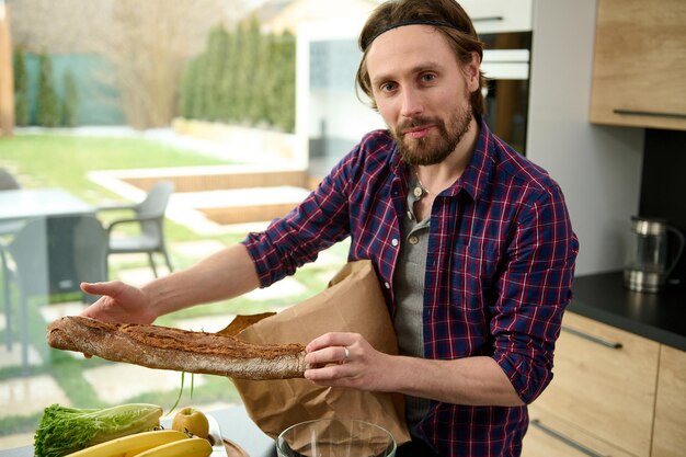 Attractive handsome young Caucasian man unpacking the eco paper bag with healthy food and showing the whole grain bread or freshly baked baguette, looking at camera standing by kitchen table at home