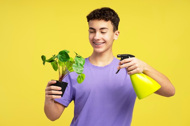 Attractive happy boy holding green flowerpot spraying it standing isolated on yellow background