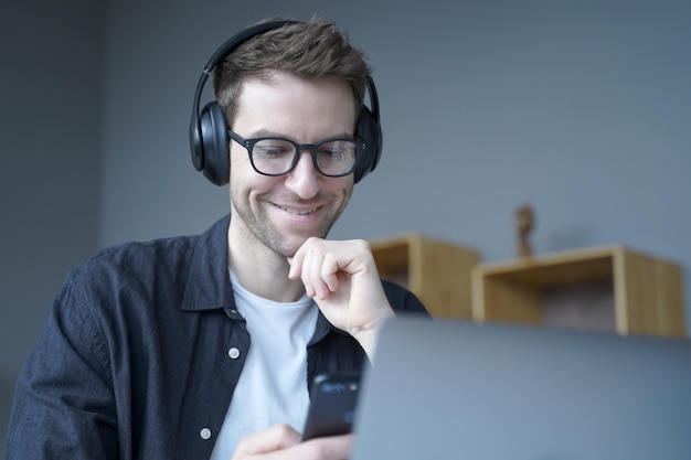 Attractive happy German home office worker in headphones and glasses sincerely smiling when looking at mobile phone screen in his hands while working remotely on laptop online Freelance concept