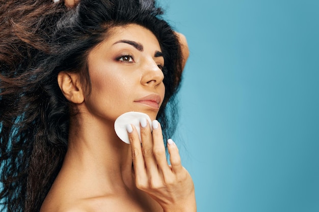 Attractive happy latin lady clean cheek with cotton pads use\
lotion toner look aside posing isolated over blue background\
cosmetic ad natural beauty skincare concept makeup removing copy\
space